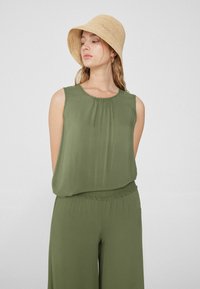 Oliwkowy top bez rękawów z plisowanym dekoltem, sparowany z luźnymi oliwkowymi spodniami. Modelka nosi słomkowy kapelusz typu bucket.