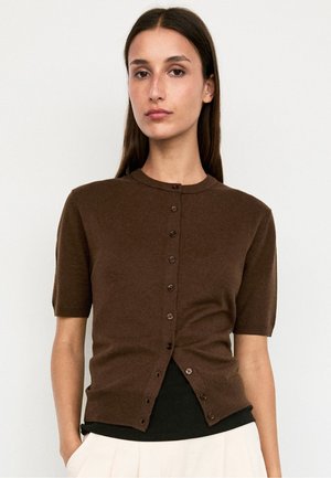Femme aux cheveux bruns raides portant un cardigan marron à manches courtes boutonné sur un top noir et un pantalon clair sur un fond uni.