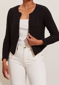 Černý pletený kardigan s texturovaným copánkovým vzorem na rukávech a výstřihu, kratšího střihu, otevřený vpředu a se dvěma předními kapsami, nošený přes bílé tričko.