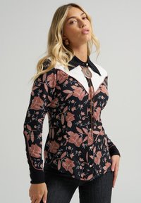 Blusa preta com padrão floral em rosa e branco, apresentando pala branca, colarinho pontudo e gravata tipo bolo decorativa. Mangas compridas com punhos de botão.