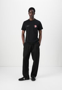 Black cotton T-shirt with logo detail, paired with loose-fitting black trousers and black sneakers, against a plain white background.