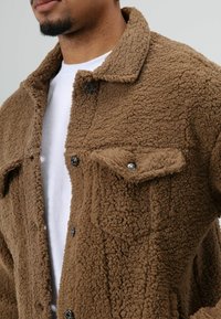 Brown, textured fleece jacket with a collar, front button closures, and two chest pockets, offering a cozy and warm appearance.