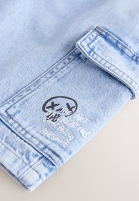 Manga de chaqueta vaquera azul claro con gráfico de calavera en blanco y negro y texto blanco ilegible en la tela cerca del puño.