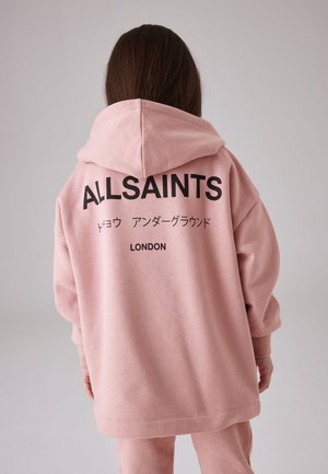 Osoba s dlhými hnedými vlasmi, ktorá má na sebe voľnú ružovú mikinu s textom "ALLSAINTS" a japonskými znakmi, stojí pred jednoduchým pozadím.