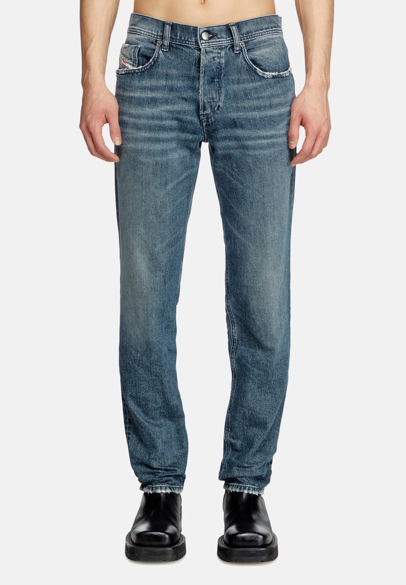 Blue denim jeans with a slim fit, featuring a faded wash, five pockets, and stitching accents. Worn with black ankle boots.
