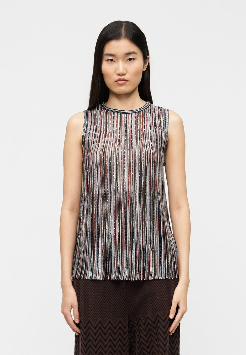 Top bez rękawów w pionowe paski w czerwonym, czarnym i srebrnym kolorze. Posiada okrągły dekolt i subtelny połysk; noszony z szerokimi brązowymi spodniami z wzorem.