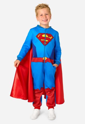 Jeune garçon souriant, portant un costume de super-héros bleu avec une cape rouge et emblème "S", tenant le bord de la cape d'une main, debout sur fond blanc.