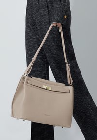 Beige læderhåndtaske med et struktureret design, der har en guldlås og kort rem. Struktur overflade og metal accenter er synlige.