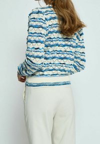 Lekki sweter z dzianiny z falistym wzorem w niebiesko-kremowe pasy, długimi rękawami i ściągaczem u dołu. Połącz z kremowymi spodniami.