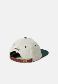 Casquette avec un corps beige et une visière verte. Présente "est. '88" brodé en noir à l'arrière, sangle en cuir marron avec attache en métal.