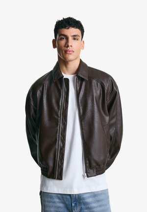 Brown leather jacket featuring a textured surface, zip front, and a classic collar. Worn over a plain white t-shirt and blue jeans.