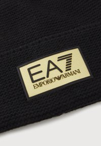 Țesătură tricotată texturată neagră, cu un eticheta care afișează "EA7 EMPORIO ARMANI" în negru pe un fundal dreptunghiular aurit.