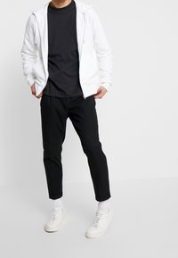 Man som bär en vit zip-up hoodie över en svart t-shirt, svarta byxor, vita strumpor och vita sneakers, står mot en enkel vit bakgrund.