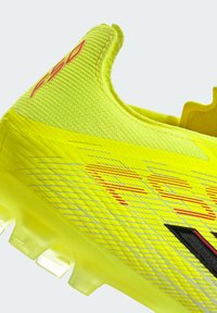 Φωτεινό κίτρινο ποδοσφαιρικό παπούτσι με υφή στην επάνω επιφάνεια, κόκκινες λεπτομέρειες και αεροδυναμικό σχεδιασμό με στρατηγικά τοποθετημένα καρφιά για καλύτερη πρόσφυση.