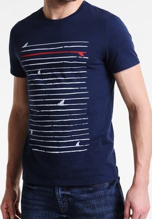 Hombre con camiseta azul marino con líneas horizontales blancas y aletas de tiburón, con una línea roja entre ellas, combinada con vaqueros azul oscuro.