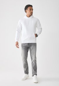 Man staat rechtop in een witte hoodie, grijze spijkerbroek, witte sneakers en een zwart horloge, kijkt naar rechts tegen een witte achtergrond.