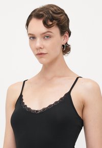 Jeune femme aux cheveux bruns courts et ondulés, portant des boucles d'oreilles en argent et un débardeur noir avec des bordures en dentelle, sur un fond blanc uni.