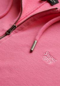 Close-up van de roze Superdry hoodie stof, met zichtbare zilveren metalen rits, trekkoordtip en geborduurd merclogo.
