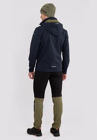 Marineblauwe softshell jas met capuchon, met een contrasterende olijfgroene voering. Zwarte broek met olijfgroene accenten en zwarte schoenen, staande tegen een effen achtergrond.