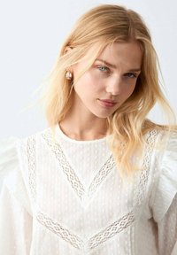 Bílá halenka s jemnými krajkovými vzory, strukturovanými puntíky a nabranými rukávy. Hladká látka s vysokým límcem a jemnými akcenty.