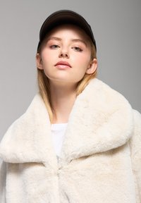 Cappotto in pelliccia sintetica crema con ampio colletto, caratterizzato da una texture morbida. Indossato sopra un top bianco e abbinato a un berretto nero, creando un look a strati.
