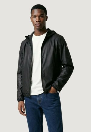 Ung mand iført en sort lynlåstrøje over en hvid skjorte og blå jeans, stående med den ene hånd i lommen mod en ensfarvet baggrund.