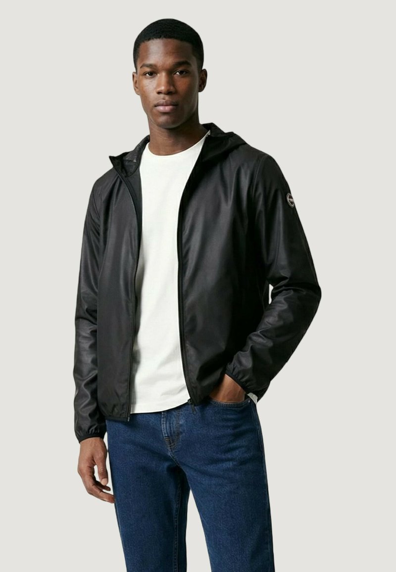 Jonge man met een zwart jack met ritssluiting over een wit shirt en een blauwe spijkerbroek, staand met één hand in zijn zak tegen een effen achtergrond.