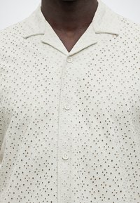 Άτομο με σκουρόχρωμο δέρμα που φοράει λευκό πουκάμισο με κουμπιά και κέντημα με παραθυράκια, με γιακά τύπου κάμπινγκ.