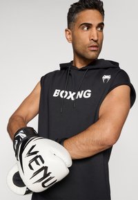 Svart ermeløs hettejakke med teksten "BOXING", båret av en person som holder hvite boksehansker med svarte detaljer og merkevare.