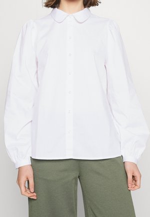 Femme portant une chemise blanche à manches longues avec col arrondi et un pantalon vert olive, debout devant un fond uni.