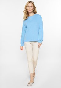 Jasnoniebieski sweter z długimi, bufiaste rękawami ozdobionymi białymi koralikami. W połączeniu z jasnobeżowymi spodniami typu slim fit i płaskim obuwiem.