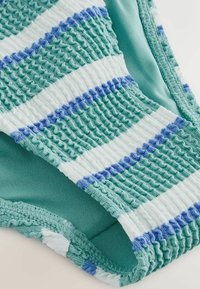 Nahaufnahme eines strukturierten Stoffs mit abwechselnden grünen und weißen Streifen, die von dünnen blauen Linien umrandet sind und Waffel- sowie glatte Strickmuster aufweisen.