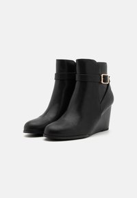 Botas de tobillo de cuero negro con tacón de cuña, que cuentan con una correa envolvente y un detalle de hebilla en tono dorado. Textura suave, diseño de punta redonda.