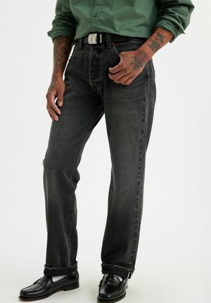 Homme portant un jean droit gris foncé, une ceinture noire, des mocassins noirs et une chemise verte boutonnée avec les manches retroussées montrant des tatouages sur les avant-bras.