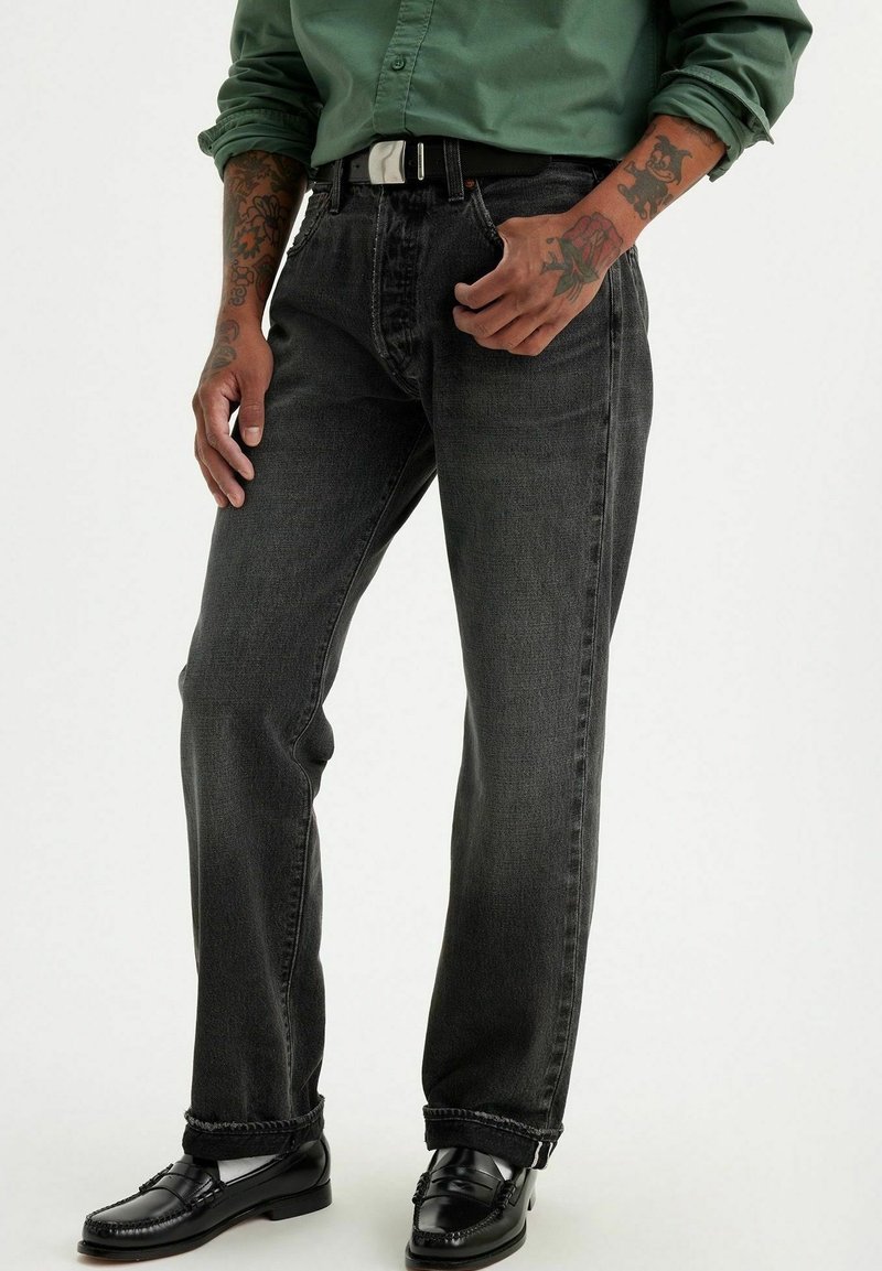 Homme portant un jean droit gris foncé, une ceinture noire, des mocassins noirs et une chemise verte boutonnée avec les manches retroussées montrant des tatouages sur les avant-bras.