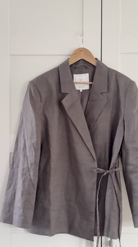 Gray linen blazer with a notched collar. Features a belt tie and textured fabric, displaying a relaxed fit and long sleeves.