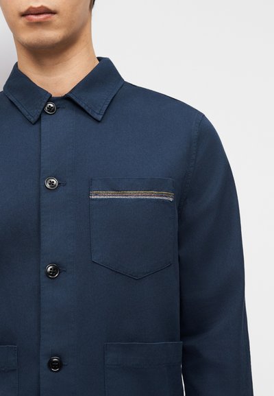 Άνδρας που φοράει μπλε σκούρο πουκάμισο με τρία μαύρα κουμπιά και τσέπη στο στήθος με πολύχρωμη οριζόντια ραφή.