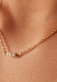 Gouden ketting met drie ongelijke witte parels, die een gestructureerd oppervlak tonen tegen een huidachtige achtergrond.