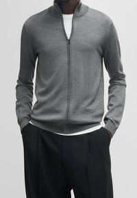 Šedá svetr na zip se stojáčkem, vyrobený z hladkého pleteného materiálu. Dlouhé rukávy a design s žebrovaným lemem, vrstvený přes bílý tričko.