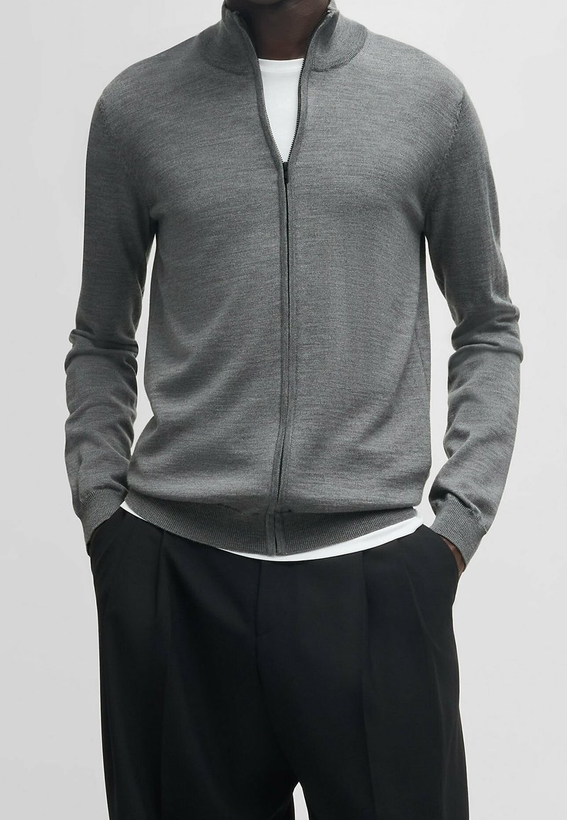 Šedá svetr na zip se stojáčkem, vyrobený z hladkého pleteného materiálu. Dlouhé rukávy a design s žebrovaným lemem, vrstvený přes bílý tričko.