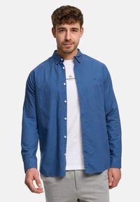 Ung man som bär en uppknäppt blå skjorta över en vit t-shirt med text, stående mot en enfärgad ljus bakgrund.