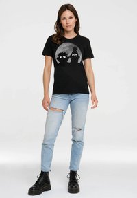 Schwarzes T-Shirt mit einem runden Grafik, die zwei stilisierte Gesichter vor einem grauen Mond zeigt. Das Modell trägt ausgewaschene hellblaue Jeans und schwarze Stiefel.