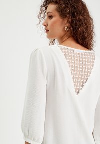 Blouse blanche à manches trois-quarts, avec un tissu texturé et un détail en dentelle à motif en losange en forme de V au niveau du dos.