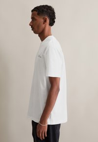 Witte korte mouwen t-shirt met een ronde hals, zijsplitten en minimalistische tekstdetail op de bovenste linkerkant van de borst. Gladde katoen materiaal.