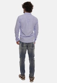 Camisa de manga larga azul claro con cuello y pliegue en la parte posterior, combinada con jeans gris deslavado y zapatillas marrones con detalles en blanco.