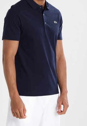 Mann trägt ein marineblaues Polohemd mit kleinem Krokodil-Logo auf der Brust, kombiniert mit weißen Shorts.