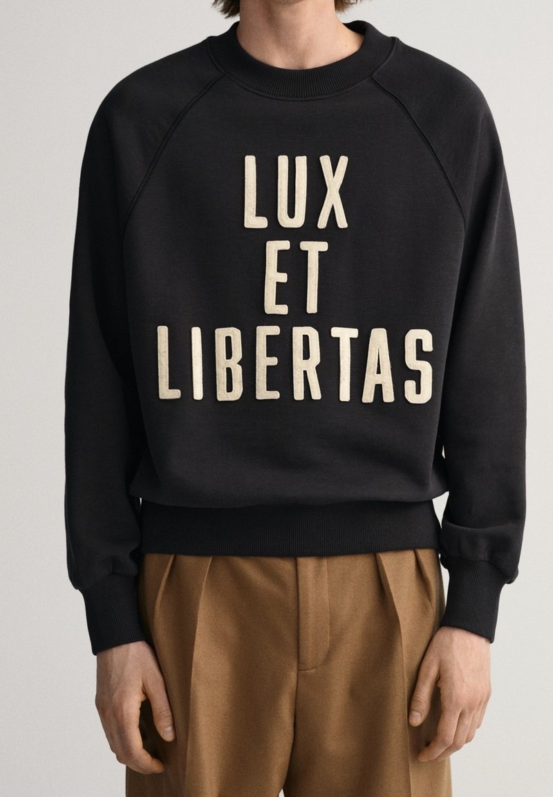 Person in einem schwarzen Sweatshirt mit großem beigem Text "LUX ET LIBERTAS" und braunen, gefalteten Hosen, der vor einem schlichten Hintergrund steht.