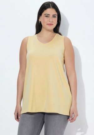 Kobieta z długimi ciemnymi włosami, ubrana w żółtą bluzkę bez rękawów i szare spodnie, stojąca na jednolitym białym tle.