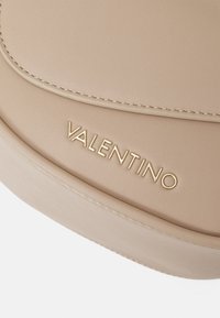 Superficie in pelle beige con texture liscia, caratterizzata da cuciture in rilievo e un logo dorato "VALENTINO" impresso. Forma arrotondata con contorni sottili.
