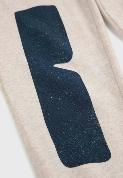 Beige Stoff mit einem großen, dunkelblauen, glitzernden Grafikelement, das dem Buchstaben 'R' ähnelt. Die Oberflächenstruktur wirkt glatt und weich.